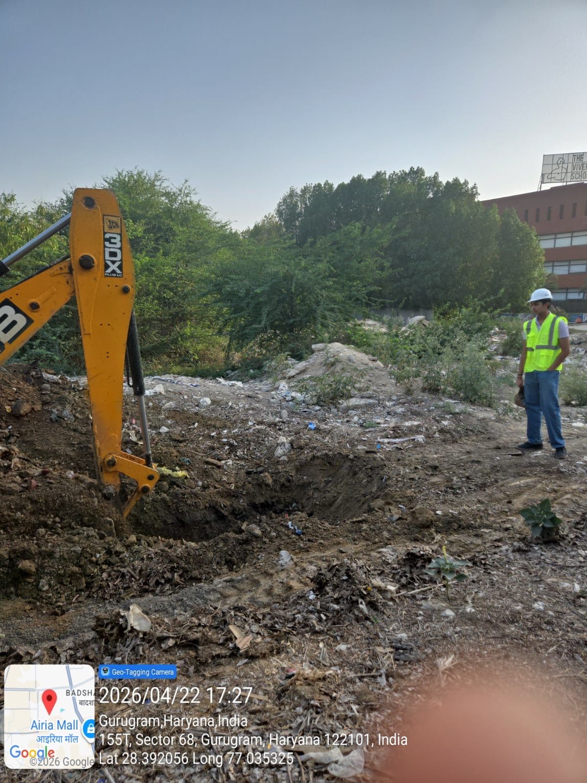 An excavator digging into littered soil while a person in a hard hat watches